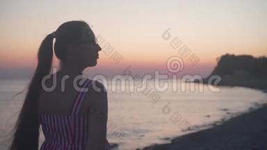 一个年轻的女孩站在海上日落的背景上。 一个女人背对着镜头站在海滩上。